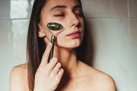 Crop concentrated young female with closed eyes massaging cheek with jade roller against ceramic wall