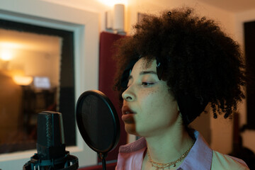 Black female singer performing song against microphone with pop filter while standing and looking forward in sound studio