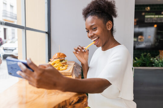 Side view joyful young African American female in white blouse enjoying delicious French fries and burger while taking selfie on smartphone in fast food restaurant