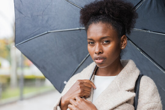 Young trendy African American female in warm coat standing with umbrella on modern city street and looking at camera