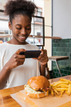 Side view calm African American female in stylish wear taking pictures of yummy burger and fries served on high table in fast food restaurant