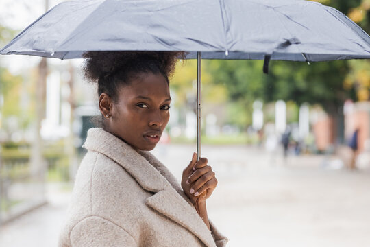 Young trendy African American female in warm coat standing with umbrella on modern city street and looking at camera