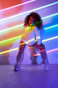 Full body stylish confident African American female dancer with curly hair and sunglasses crouched looking at camera in neon lights in dancing studio