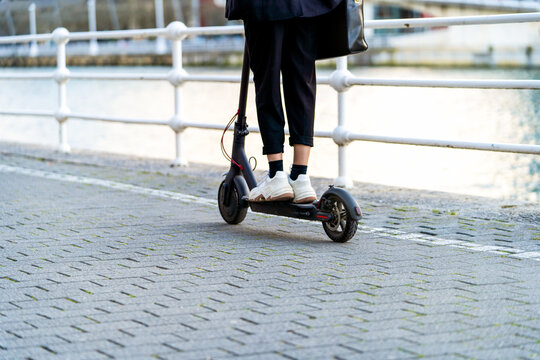 Side view of crop anonymous female in sneakers riding modern scooter on footpath against city river