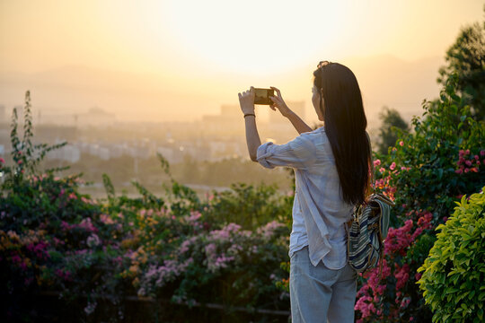 Ethnic Asian woman standing in garden taking picture with smartphone during sunset time