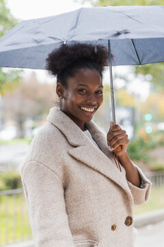 Young trendy African American female in warm coat standing with umbrella on modern city street and looking at camera