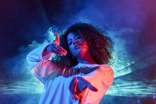 Portrait of energetic young African American female in white wear outstretching arms towards camera while dancing in dark studio in neon lights