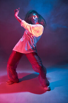 Full length fit young African American female dancer in loose informal wear looking at camera while dancing in dark studio in neon lights