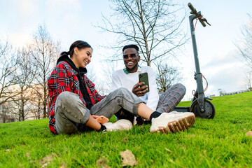 From below of cheerful multiracial couple in trendy wear with cellphone speaking on meadow with electric scooter in daytime