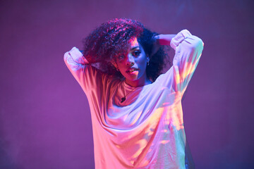 Portrait of young African American female dancer in loose informal wear touching curly hair and looking at camera while dancing in dark studio in neon lights