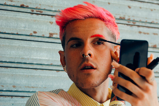 Homosexual man with piercings and modern haircut applying mascara on eyelashes with applicator against cellphone