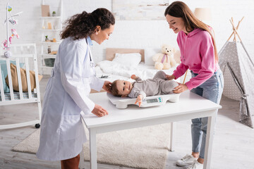 happy african american pediatrician and mother looking at infant boy on baby scale