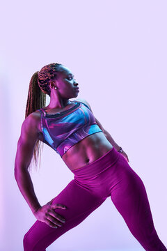 Young muscular African American female athlete in sports clothes with Afro braids and hands on hips looking away in neon light