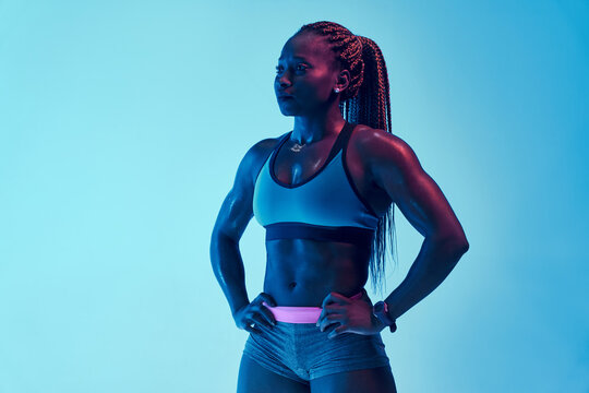 Young muscular African American female athlete in sports clothes with Afro braids and hands on hips looking away in neon light