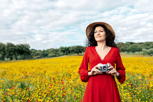 Smiling female in hat taking photo on vintage camera on meadow under cloudy sky