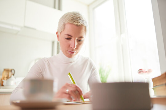 Smiling female astrologist taking notes in notepad at desk with cup of coffee at home in sunlight