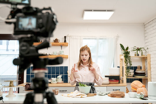 Young female blogger talking against smartphone while recording culinary video and looking down at home