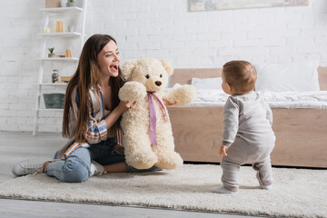 amazed young mother holding teddy bear near infant son