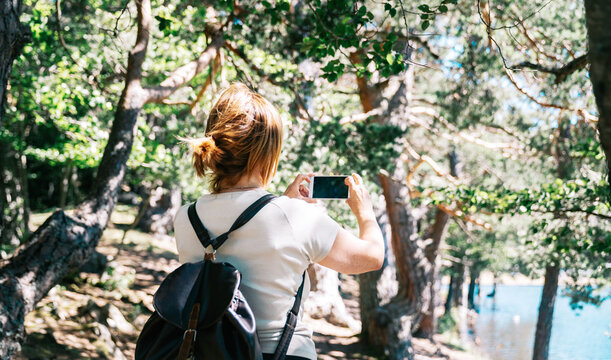 Back view of anonymous female backpacker taking photo of lake on cellphone in Aran Valley of Lleida Catalonia Spain