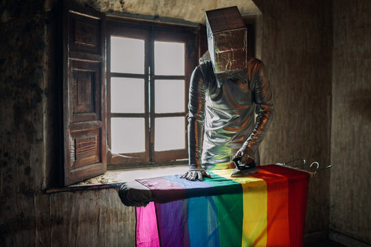Unrecognizable person in silver costume and box on head ironing LGBTQ flag while standing in shabby abandoned room