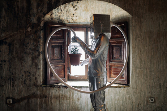 Side view unrecognizable person wearing silver costume with breathing apparatus and hose attached to potted plant standing in shabby abandoned room