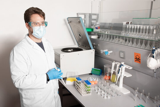 Male chemist in uniform and disposable mask looking forward at table with professional equipment and test tubes in laboratory