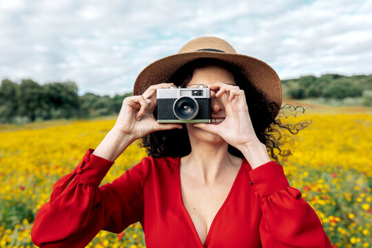 Anonymous smiling female in hat taking photo on vintage camera on meadow under cloudy sky