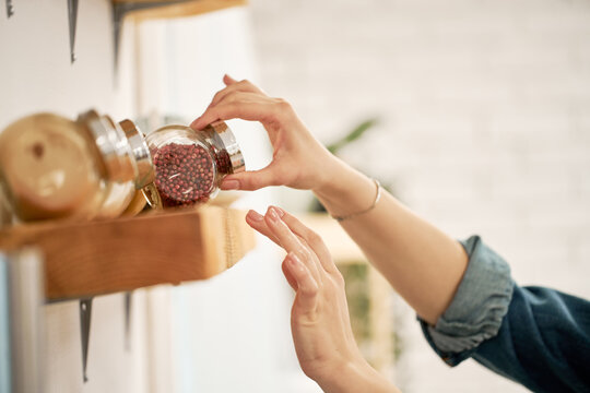 Crop unrecognizable female taking glass jar of red peppercorns from wooden shelf with assorted spices in house