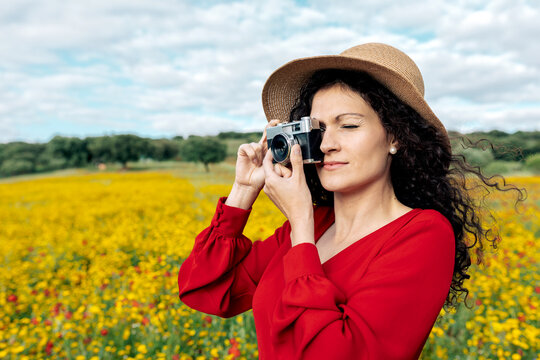 Smiling female in hat taking photo on vintage camera on meadow under cloudy sky