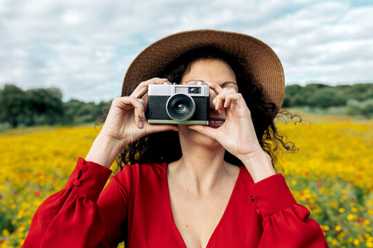 Anonymous smiling female in hat taking photo on vintage camera on meadow under cloudy sky