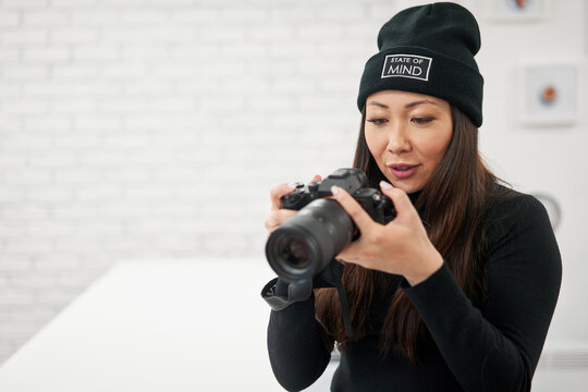 Ethnic female in black beanie and shirt using digital photo camera on blurred background