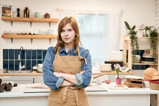 Young self assured female with folded arms in apron looking at camera at home