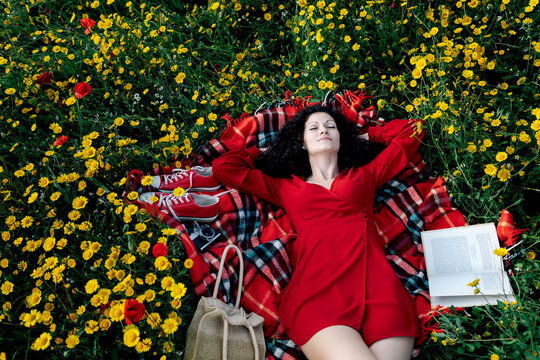 From above of female with eyes closed having a break from reading textbook on blanket with handbag among blossoming daisies during a spring summer day