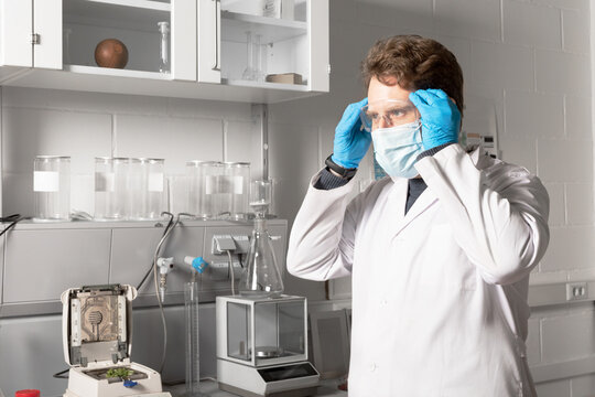Male chemist in sterile mask and gloves looking forward against moisture analyzer and analytical balance in cannabis laboratory