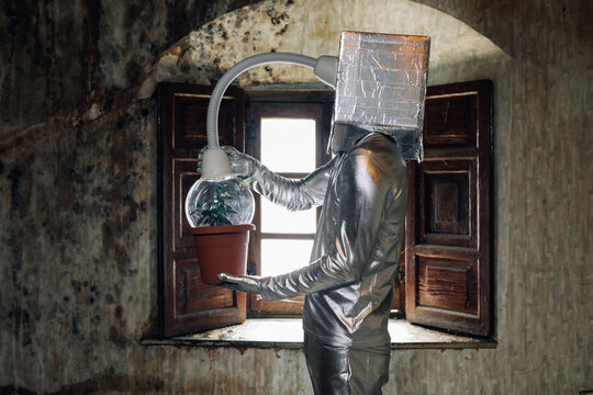 Side view unrecognizable person wearing silver costume with breathing apparatus and hose attached to potted plant standing in shabby abandoned room