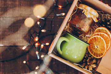 Gift box with tea set. Tea, cup, jam or honey, spices, dried oranges, anise in box on wooden table with christmas lights, top view. Beautiful  present, care package for family and friends