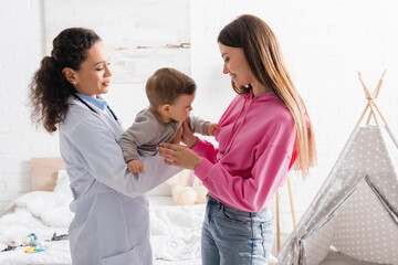 Fototapeta premium smiling african american pediatrician in white coat holding infant boy near happy mother