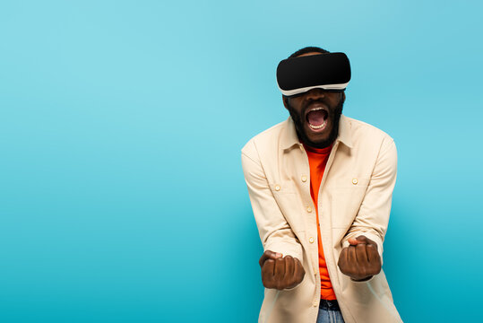 angry african american man in vr headset screaming and showing clenched fists isolated on blue