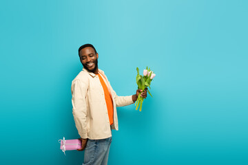 cheerful african american man holding tulips and while hiding present behind back on blue