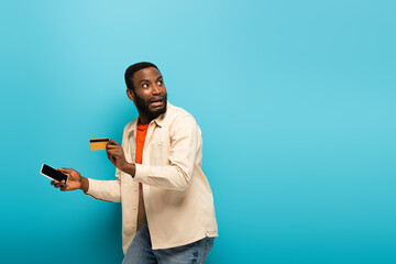 scared african american man with mobile phone and credit card looking away on blue