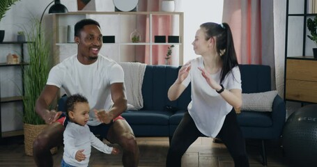 Likable smiling active fit modern diverse married couple squating together during family home workout near their playful cute little child