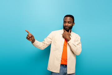 thoughtful african american man touching face and pointing away with finger on blue background