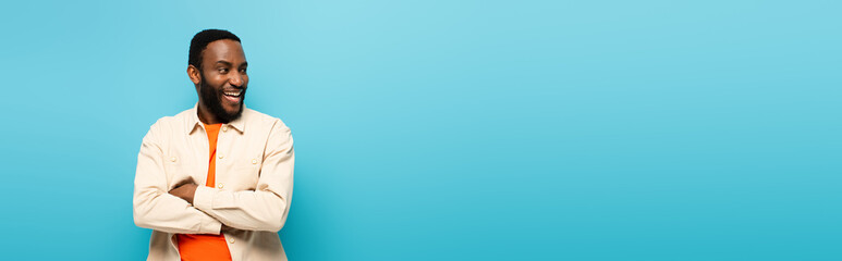 excited african american man with crossed arms laughing while looking away isolated on blue, banner