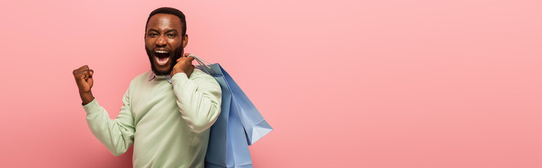 excited african american man with shopping bags showing yeah gesture isolated on pink, banner