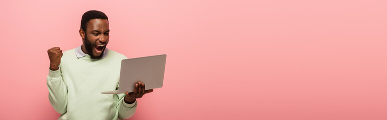 excited african american man showing success gesture while holding laptop isolated on pink, banner