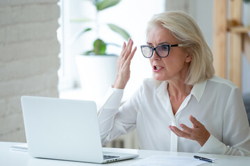 Angry senior female employee work on laptop in office frustrated by slow internet connection on...