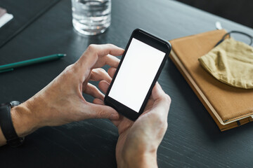 Male hands holding smartphone with blank screen at table with organizer, cloth mask and pen