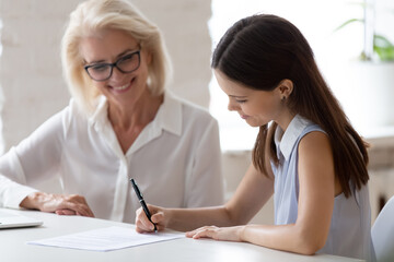 Smiling young Caucasian female employee put signature on contract close deal with middle-aged employer in office. Happy diverse businesswoman sign document make agreement after successful meeting.