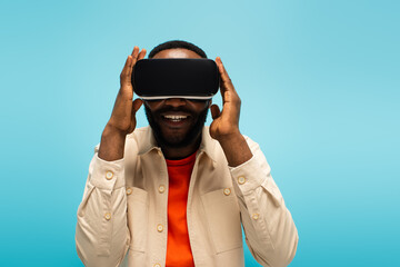 cheerful african american man adjusting vr headset while gaming isolated on blue