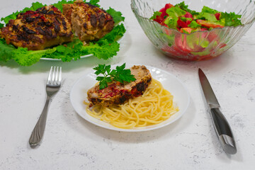 A plate of pasta and a large piece of pork cooked in the oven, baked in the oven. Oven baked meat on green lettuce leaves. With pasta on a plate with a fork and a knife. Against the background of a